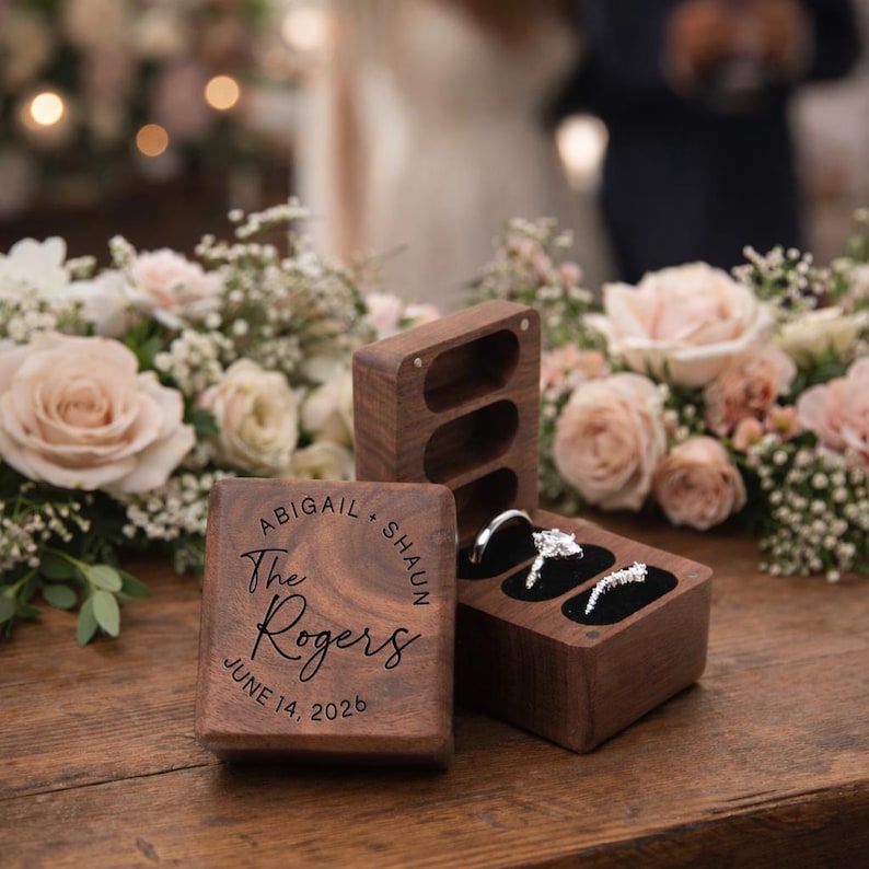 May include: Wooden ring boxes with a wedding theme. One box is engraved with "Abigail + Shaun The Rogers JUNE 14, 2026". The other box holds two rings. The boxes are on a wooden surface with flowers in the background.