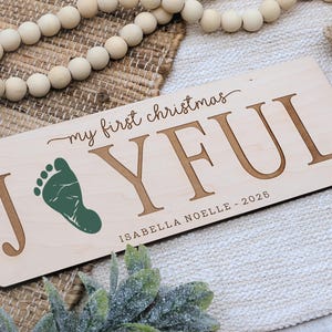 May include: A rectangular wooden sign with the words "my first christmas JOYFUL" in brown lettering. A green footprint is next to the letter "J". The name "ISABELLA NOELLE - 2026" is below. The sign is on a textured surface.