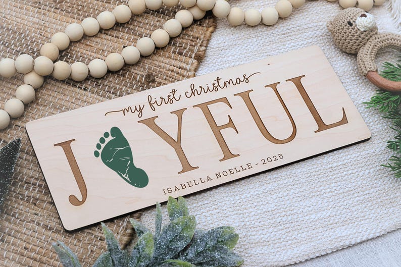 May include: A rectangular wooden sign with the words "my first christmas JOYFUL" in brown lettering. A green footprint is next to the letter "J". The name "ISABELLA NOELLE - 2026" is below. The sign is on a textured surface.