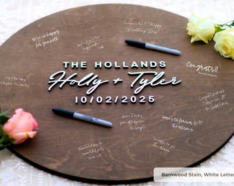 May include: A round wooden wedding guest book with the names "Holly + Tyler" and the date "10/02/2025" in white lettering. The wooden surface is stained brown. Two black markers and floral accents are included.