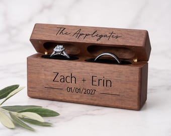May include: A rectangular dark wood ring box with a hinged lid, featuring the engraved text The Applegates on the lid and Zach + Erin 01/01/2027 on the front. The open box contains two black velvet slots holding a diamond ring and a plain metal band.