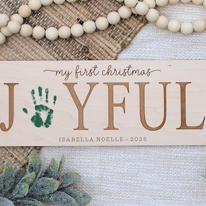May include: A rectangular wooden sign with the words "my first christmas JOYFUL" in a script font and block letters. A green handprint is on the left. The name "ISABELLA NOELLE - 2025" is below. The sign is surrounded by Christmas decorations.
