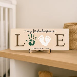 May include: A wooden sign with the words "my first christmas" in cursive. The word "LOVE" is spelled out with the letters L and E, with a green handprint and white footprint in between. The name "LIAM NOAH - 2025" is printed below.
