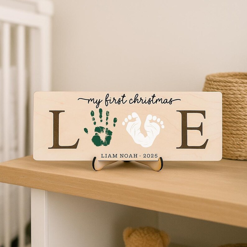 May include: A wooden sign with the words "my first christmas" in cursive. The word "LOVE" is spelled out with the letters L and E, with a green handprint and white footprint in between. The name "LIAM NOAH - 2025" is printed below.