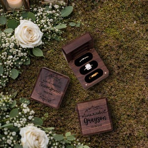 May include: Three wooden ring boxes, two closed and one open, displaying engagement rings. The open box reveals three rings: a diamond band, a square-cut diamond ring, and a gold band. The closed boxes are engraved with names and dates.