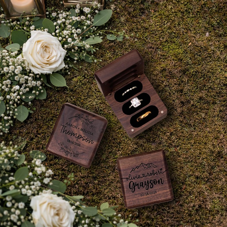 May include: Three wooden ring boxes, two closed and one open, displaying engagement rings. The open box reveals three rings: a diamond band, a square-cut diamond ring, and a gold band. The closed boxes are engraved with names and dates.