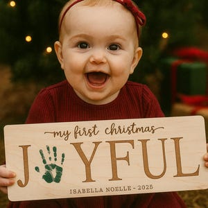 May include: A smiling baby wearing a red headband and dress holds a wooden sign that says "my first christmas JOYFUL" with a green handprint and the name "Isabella Noelle - 2025". The background includes a Christmas tree and blurred lights.