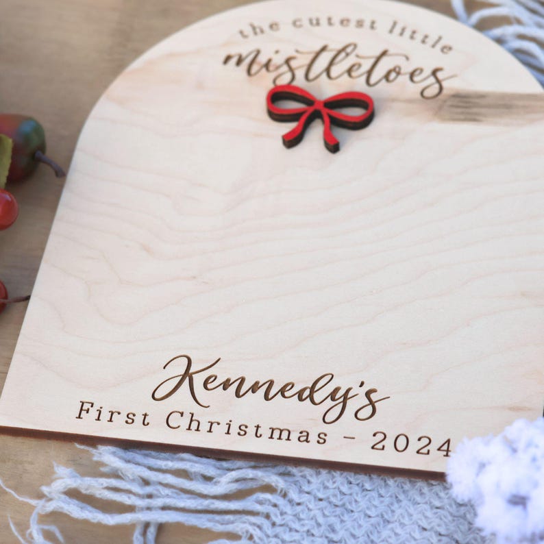 May include: A wooden sign with the text "the cutest little mistletoes" and a red bow. The sign also has the text "Kennedy's First Christmas - 2024"