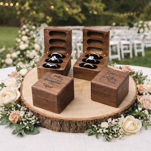 May include: Four wooden ring boxes, two open and two closed, sit on a wooden slice surrounded by flowers. The open boxes display rings on black velvet. The closed boxes are engraved with names and a date. Wedding theme.