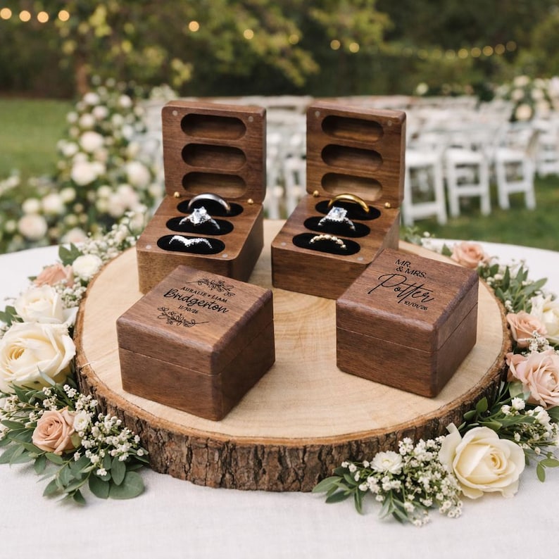 May include: Four wooden ring boxes, two open and two closed, sit on a wooden slice surrounded by flowers. The open boxes display rings on black velvet. The closed boxes are engraved with names and a date. Wedding theme.