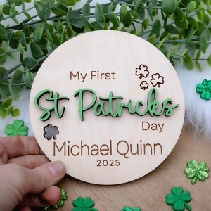 以下が含まれることがあります： 緑と茶色の文字で「My First St. Patricks Day」と書かれた丸い木製の看板。看板には「Michael Quinn」という名前と「2025」という年号も書かれている。