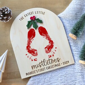 May include: A wooden sign with the text "The Cutest Little Mistletoes Maddie's First Christmas - 2024" and two red handprints. A green holly leaf with a red berry is above the handprints.