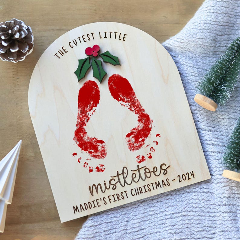 May include: A wooden sign with the text "The Cutest Little Mistletoes Maddie's First Christmas - 2024" and two red handprints. A green holly leaf with a red berry is above the handprints.