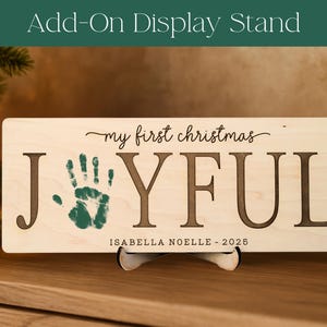 May include: A rectangular wooden sign with the words "my first christmas JOYFUL" and a green handprint. The sign also includes the name "ISABELLA NOELLE - 2026". The sign is displayed on a stand, with a Christmas tree and ornaments in the background.