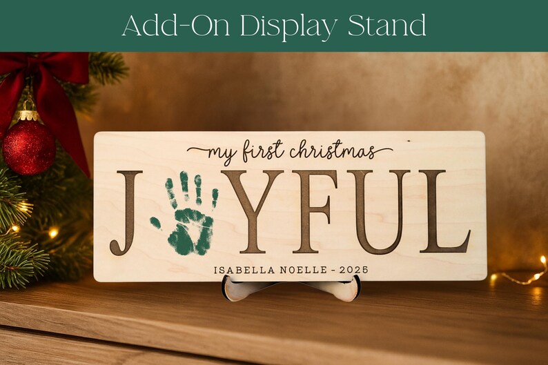 May include: A rectangular wooden sign with the words "my first christmas JOYFUL" and a green handprint. The sign also includes the name "ISABELLA NOELLE - 2026". The sign is displayed on a stand, with a Christmas tree and ornaments in the background.