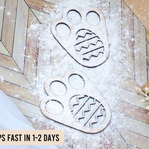 May include: Two wooden paw print cutouts with Easter egg designs, dusted with white powder, rest on a light brown, chevron-patterned surface. Decorative foliage and a white cloth add to the Easter theme.