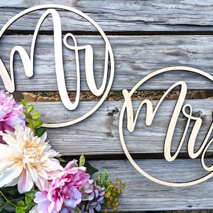 May include: Two wooden circles with the words "Mr." and "Mrs." written in cursive script. The circles are on a wooden background with a floral arrangement in the bottom left corner.