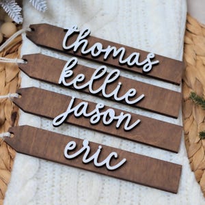May include: Four wooden name tags with white cursive names: Thomas, Kellie, Jason, and Eric. The tags are dark brown with a hole for string. The tags are on a white knitted surface.