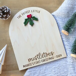 May include: A wooden sign with the text "The Cutest Little Mistletoes Maddie's First Christmas - 2024" and a green holly leaf with red berries.