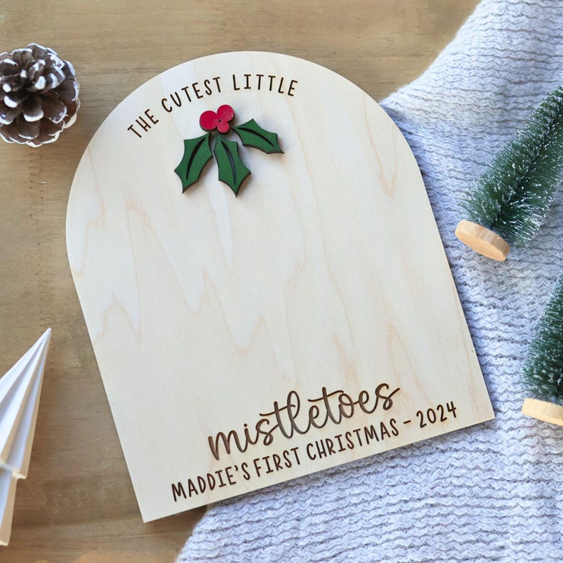May include: A wooden sign with the text "The Cutest Little Mistletoes Maddie's First Christmas - 2024" and a green holly leaf with red berries.
