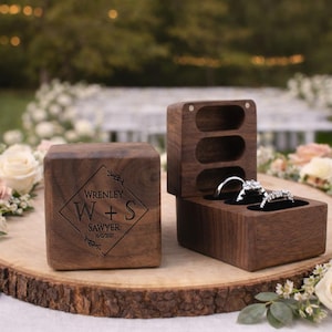 May include: Two wooden ring boxes, one closed and one open, displaying two diamond rings. The closed box is engraved with "Wrenley W+S Sawyer 10/12/2021". The boxes are on a wooden slice, with a blurred floral background.