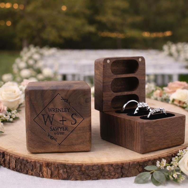 May include: Two wooden ring boxes, one closed and one open, displaying two diamond rings. The closed box is engraved with "Wrenley W+S Sawyer 10/12/2021". The boxes are on a wooden slice, with a blurred floral background.