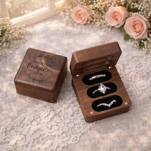 May include: A wooden ring box with three slots, one containing a silver band, another a diamond ring, and the third a curved band. The box is open, revealing the rings. The closed box has the names "AURALIE + LIAM" and "BRIDGERTON" with the date "11/30/26" engraved on the lid.