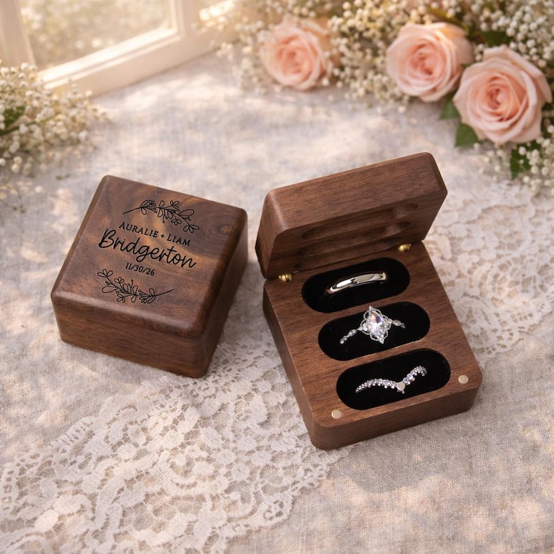May include: A wooden ring box with three slots, one containing a silver band, another a diamond ring, and the third a curved band. The box is open, revealing the rings. The closed box has the names "AURALIE + LIAM" and "BRIDGERTON" with the date "11/30/26" engraved on the lid.
