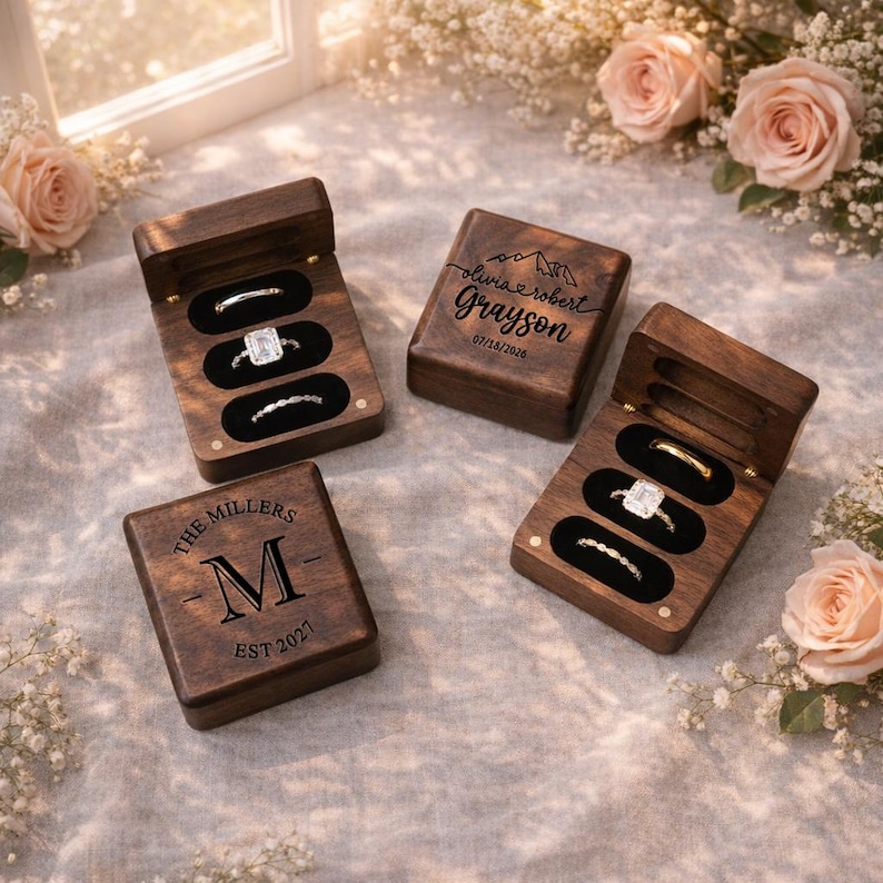 May include: Four wooden ring boxes, two open to display rings. The boxes are engraved with names, dates, and initials. The rings include a gold band, a diamond solitaire, and other delicate bands. The boxes are surrounded by flowers.