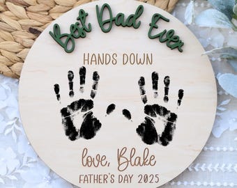 Puede incluir: Un letrero redondo de madera con el texto "Best Dad Ever" en letras verdes, "Hands Down" en letras marrones y "love, Blake" en letras marrones. El letrero también tiene dos huellas de manos negras y el texto "Father's Day 2025" en letras marrones.