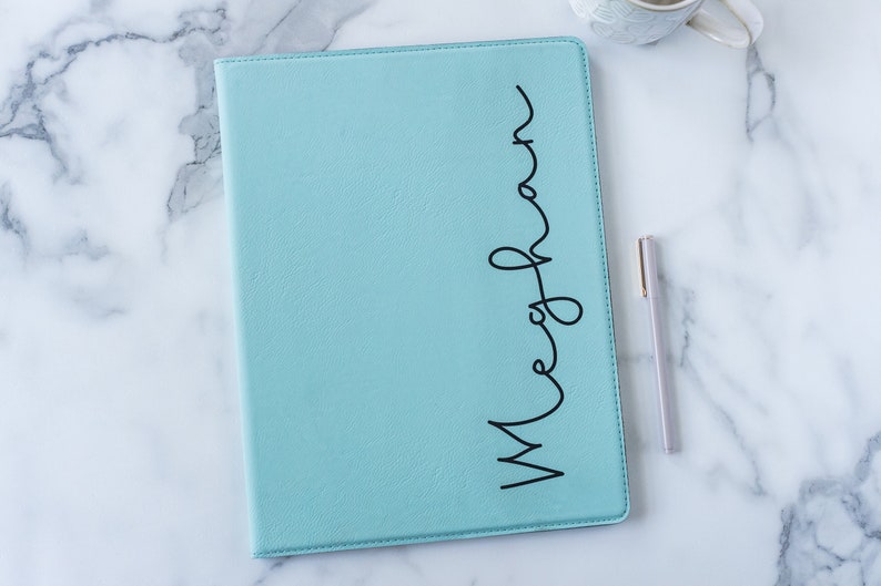 May include: A light blue leather-bound journal with the name "Meghan" written in black cursive on the front cover. A silver pen is lying on a white marble surface next to the journal.