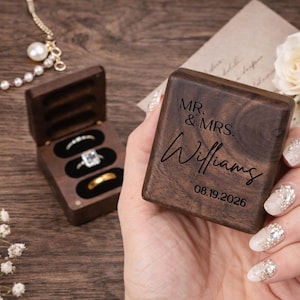 May include: A wooden ring box with the inscription "MR. & MRS. Williams" and the date "08.19.2026" is held in a hand. The box is open, revealing three ring slots with rings. A pearl necklace and white flowers are also visible.