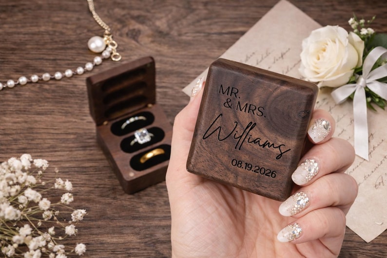 May include: A wooden ring box with the inscription "MR. & MRS. Williams" and the date "08.19.2026" is held in a hand. The box is open, revealing three ring slots with rings. A pearl necklace and white flowers are also visible.