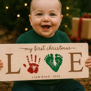 May include: A smiling baby holds a wooden sign that says "my first christmas LOVE" with a red handprint and green footprints. The sign also includes the name "LIAM NOAH - 2025". The baby is wearing a green shirt and blue jeans.