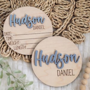 May include: Two circular wooden baby announcement plaques. The name "Hudson" is in blue script, with "Daniel" below in a smaller font. One plaque has lines for date, time, weight, and length. The plaques are on a woven mat.