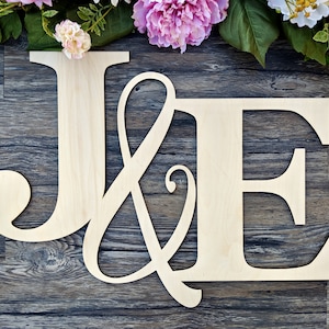 Puede incluir: Letras de madera recortadas que deletrean "J & E" en una fuente cursiva. Las letras son de color marrón claro y están sobre un fondo de madera oscura.