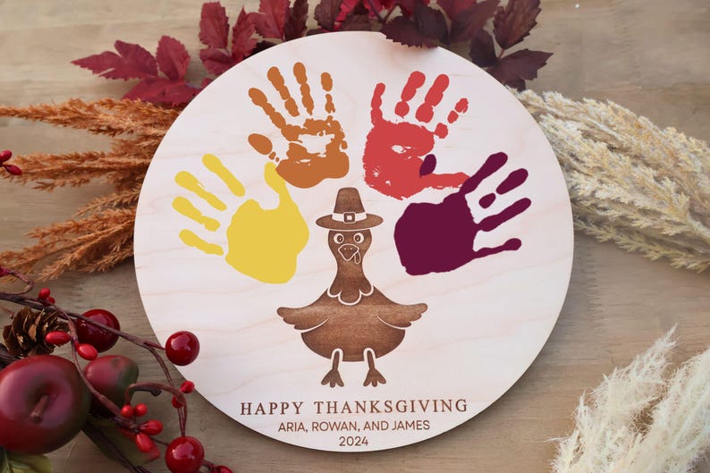 Pode incluir: Um sinal redondo de madeira branca com uma silhueta de peru no centro. Quatro impress&otilde;es de m&atilde;os em vermelho, laranja, amarelo e roxo cercam o peru. O texto "HAPPY THANKSGIVING" est&aacute; na parte inferior do sinal, seguido de "ARIA, ROWAN, AND JAMES" e "2024".