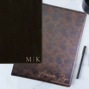 May include: Two black leather folders with gold stitching. The folder on the left has the initials "M|K" embossed in gold. The folder on the right has the name "Andrew Pope" and the title "Executive Assistant" embossed in gold.
