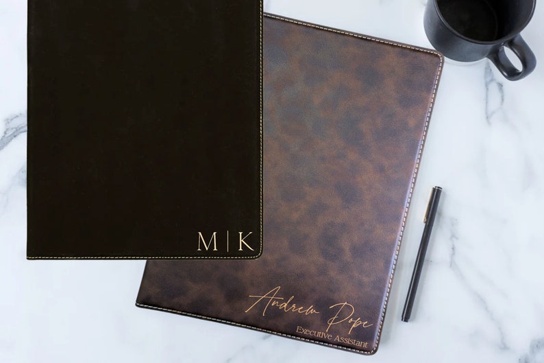 May include: Two black leather folders with gold stitching. The folder on the left has the initials "M|K" embossed in gold. The folder on the right has the name "Andrew Pope" and the title "Executive Assistant" embossed in gold.