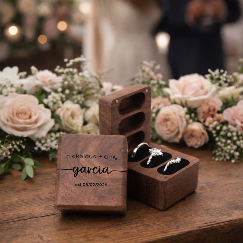 May include: Wooden ring boxes with a wedding theme. One box is open, displaying two diamond rings. The other box is closed, with the names "nickolaus + amy garcia" and the date "est 08/02/2026" engraved on the front.
