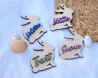 Op de afbeelding: Vier houten bunny-vormige ornamenten met namen erop geschreven in verschillende kleuren. De namen zijn Katie, Kolton, Susan en Trent.