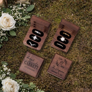 May include: Four wooden ring boxes with black velvet interiors, each holding rings. Two boxes are open, displaying engagement rings. Two closed boxes are engraved with names and dates. The background is moss and flowers.