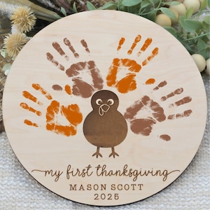 May include: A round wooden sign with a turkey design made of handprints in brown and orange. The text "my first thanksgiving" is written in cursive, with the name "MASON SCOTT" and the year "2025" below.