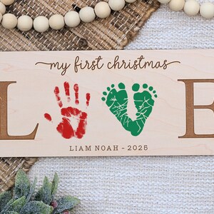 May include: Wooden sign with the words "my first christmas" and the word "LOVE" spelled out with brown letters. The "O" is a green footprint design, and the "V" is a red handprint. The name "Liam Noah" and the year "2025" are below.