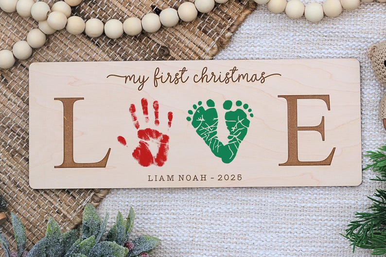 May include: Wooden sign with the words "my first christmas" and the word "LOVE" spelled out with brown letters. The "O" is a green footprint design, and the "V" is a red handprint. The name "Liam Noah" and the year "2025" are below.