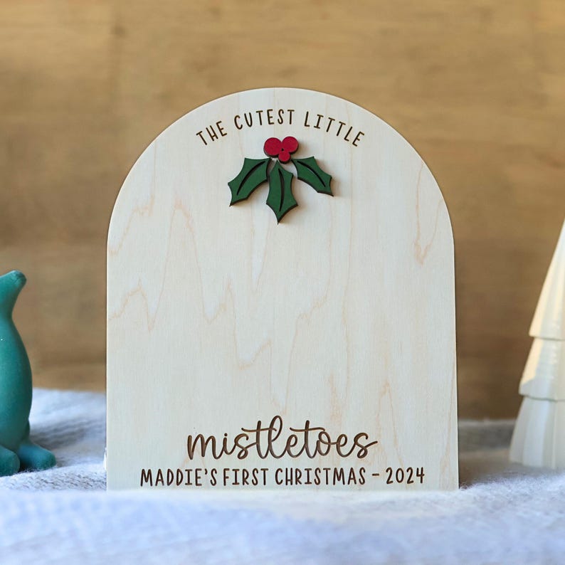 May include: A wooden sign with a rounded top, featuring the text "The Cutest Little Mistletoes Maddie's First Christmas - 2024". A green holly leaf with a red berry is attached to the top of the sign.