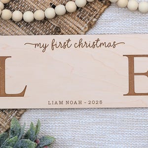 May include: A light-colored wooden sign with the words "my first christmas" in cursive script. Large brown letters "L" and "E" flank the center. The name "LIAM NOAH - 2025" is printed below. The sign is surrounded by decorative elements.