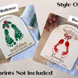 May include: Two wooden signs with the text "the cutest little mistletoes" and a pair of footprints. One sign has a green mistletoe with red berries and the other has a red bow. The text "Maddie's First Christmas - 2024" and "Kennedy's First Christmas - 2024" are on the signs.