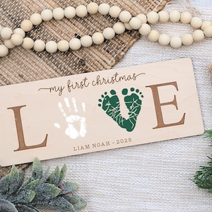 May include: A wooden sign with the words "my first christmas" and the word "LOVE" spelled out with the letters L and E in a dark brown color. The O is represented by green footprints and a white handprint. The name "Liam Noah - 2028" is below the word "LOVE".