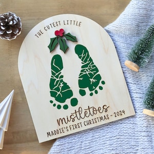 K&ouml;nnte beinhalten: Ein Holzschild mit dem Text "The Cutest Little Mistletoes Maddie's First Christmas - 2024". Das Schild zeigt zwei gr&uuml;ne Fu&szlig;abdr&uuml;cke und ein gr&uuml;nes Blatt mit einer roten Beere.